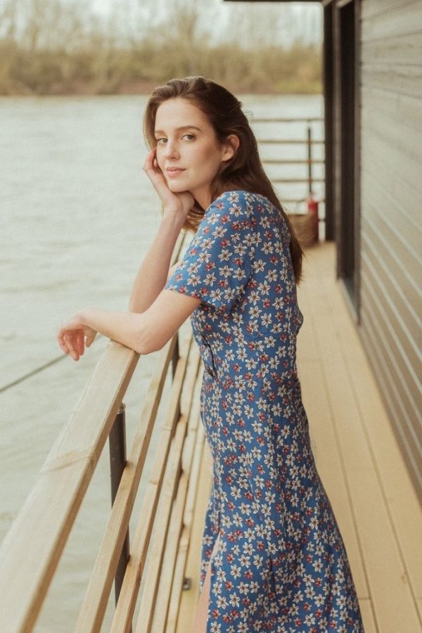 Porté de profil de la robe midi CLARISSE sur le pont d'un bateau. Le mannequin est accoudé à la balustrade et regarde l'objectif.