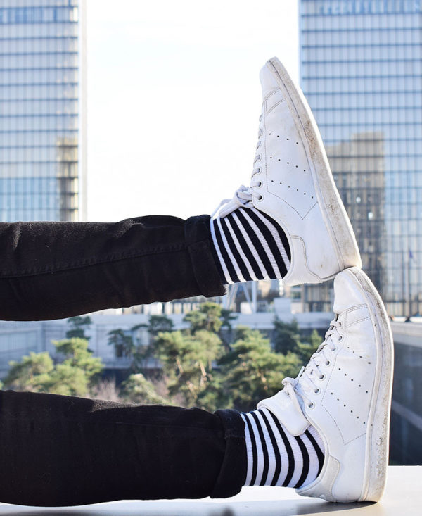 Porté des chaussettes Made in France rayées avec sneakers sur fond de building. Les deux pieds sont visibles, l'un marine rayé blanc, l'autre blanc rayé marine.