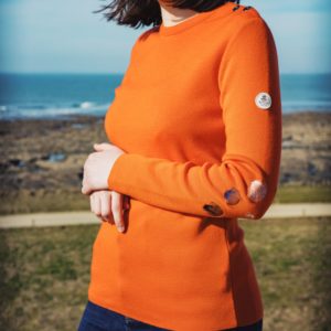 Porté du pull femme grande taille ROZEN Orange de trois quart sur fond de mer. Les broderies coquillages au coude sont visibles.