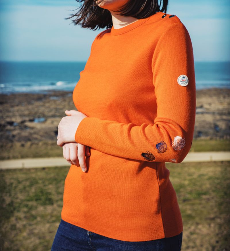 Porté du pull femme grande taille ROZEN Orange de trois quart sur fond de mer. Les broderies coquillages au coude sont visibles.
